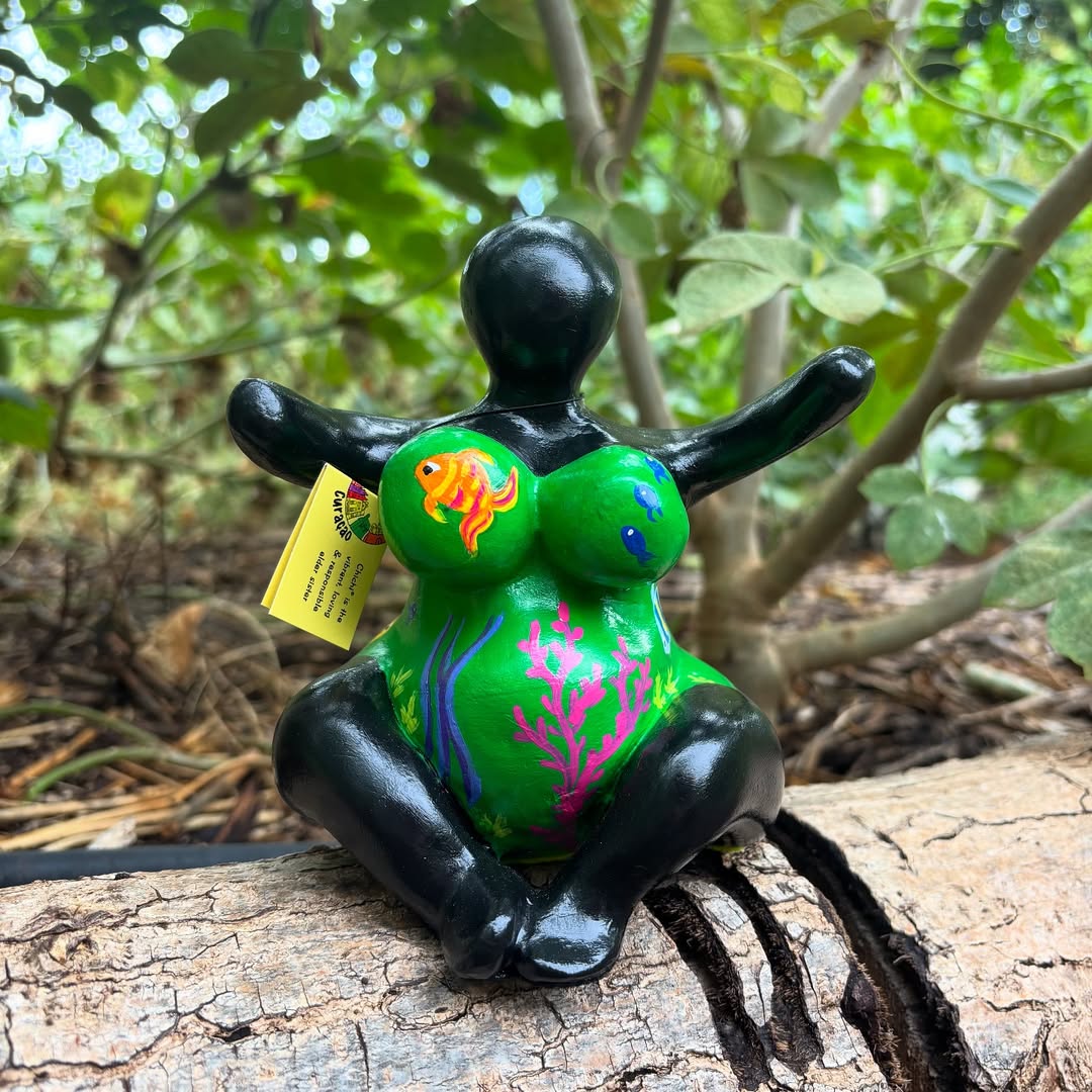 Handbeschilderd Chichi Curaçao beeldje met groene en zwarte kleuren, zittend op een boomstam in een natuurlijke omgeving.