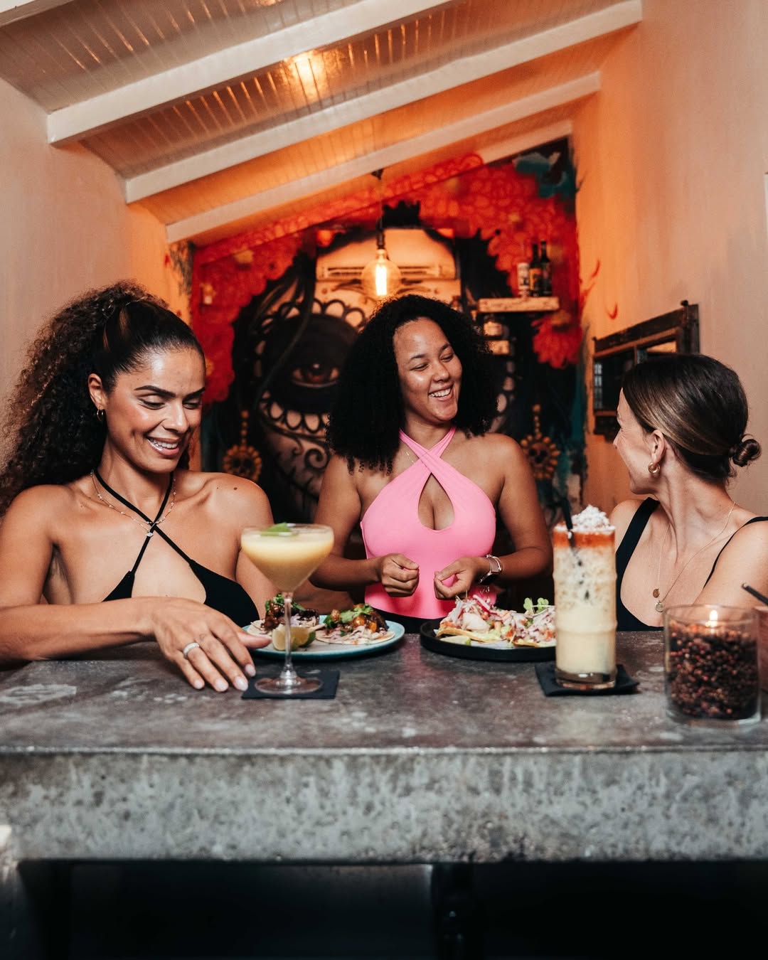 Drie vrouwen genieten van cocktails en gedeelde tapas bij Mosa Cana Bar & Kitchen Curaçao in een kleurrijke en artistieke setting